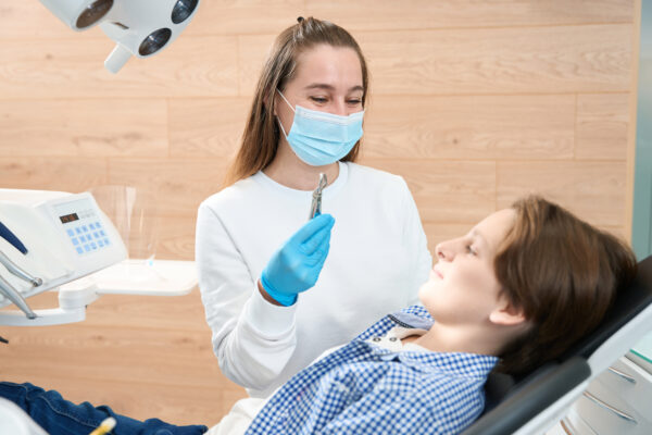 Smiling female dentist showing a boy a special tool for tooth extraction, she is wearing gloves and a mask