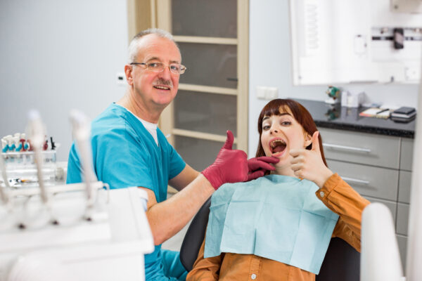 Beautiful young woman at dentist's office satisfied after successful dental treatment, sitting in the chair and showing thumb up together with senior male doctor dentist.