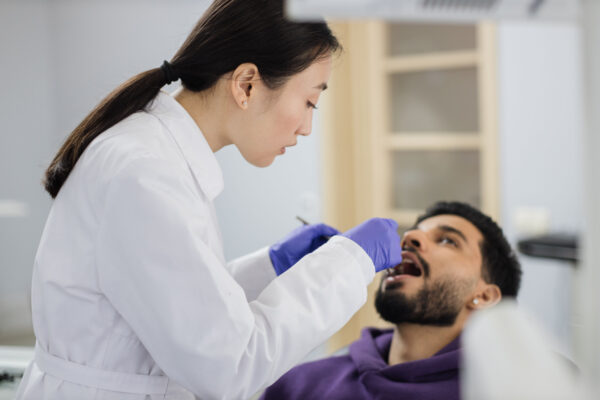 Beautiful smile with white teeth. Asian dentist examines the oral cavity of young bearded handsome man through a magnifying glass in the dental office clinic.