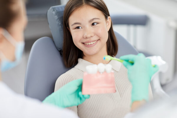 Portrait of smiling Asian girl sitting in dental chair and listening to dentist holding tooth model, copy space